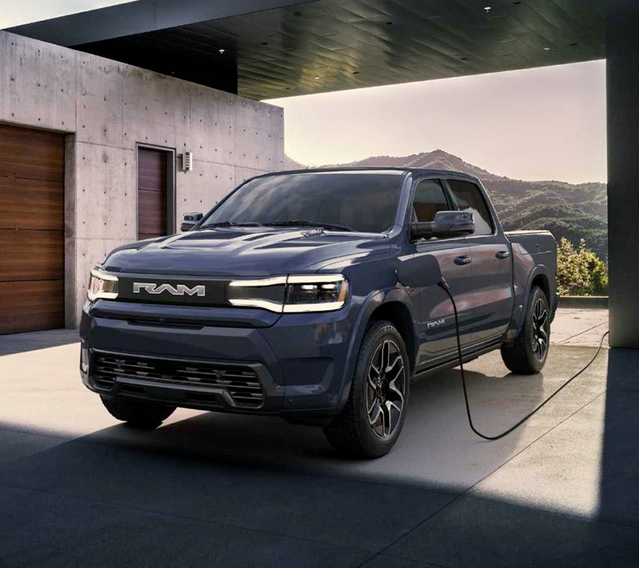 The Ram 1500 Rev Crew Cab parked in the driveway of a building with a charger cord plugged into its charging port.