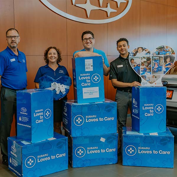 Four workers with charity boxes at Subaru dealership.