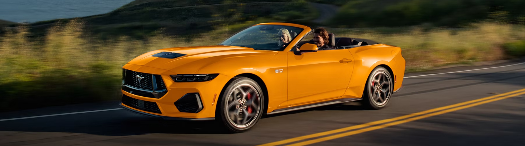 Two people in a 2026 Ford Mustang® GT convertible in Orange Fury Metallic Tri-coat are being driven on a coastal highway