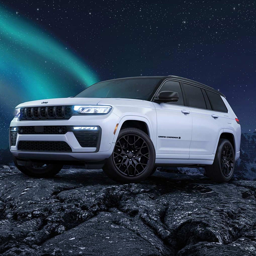 A white and black 2027 Jeep Grand Cherokee Summit Obsidian, parked on a rocky hill at night, with stars and a trail of the northern lights in the background.