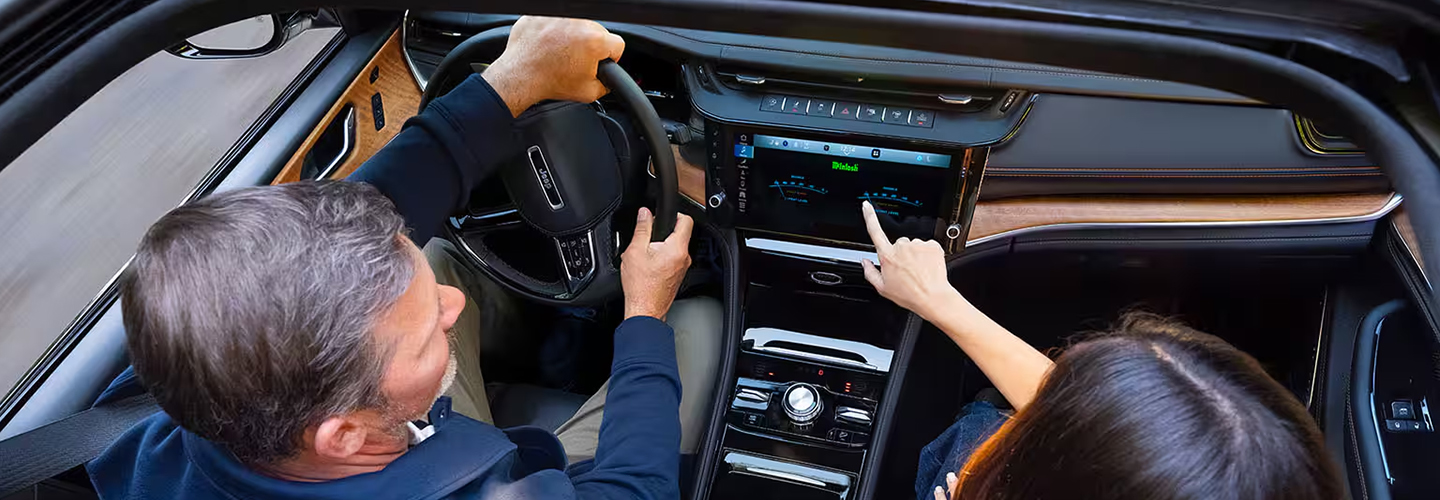 The front seat area of the 2027 Jeep Grand Cherokee Summit, with a man in the driver seat and a woman in the passenger seat. Shot from outside the open sunroof.