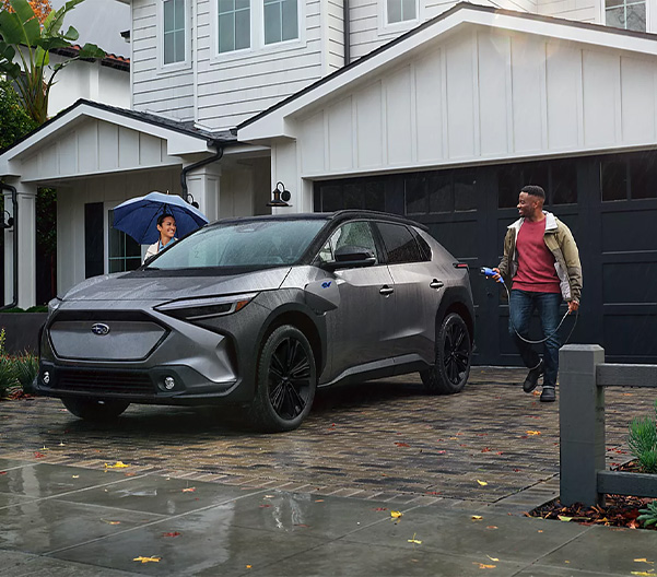 A couple walks from a white house toward a blue Subaru Solterra AWD electric SUV.